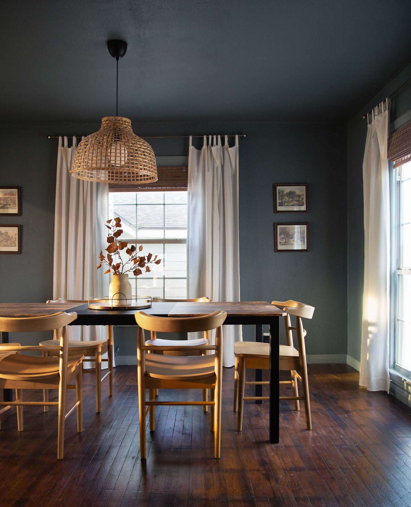 Dining room with wooden table and chairs, wicker pendant light, and large window with white curtains.