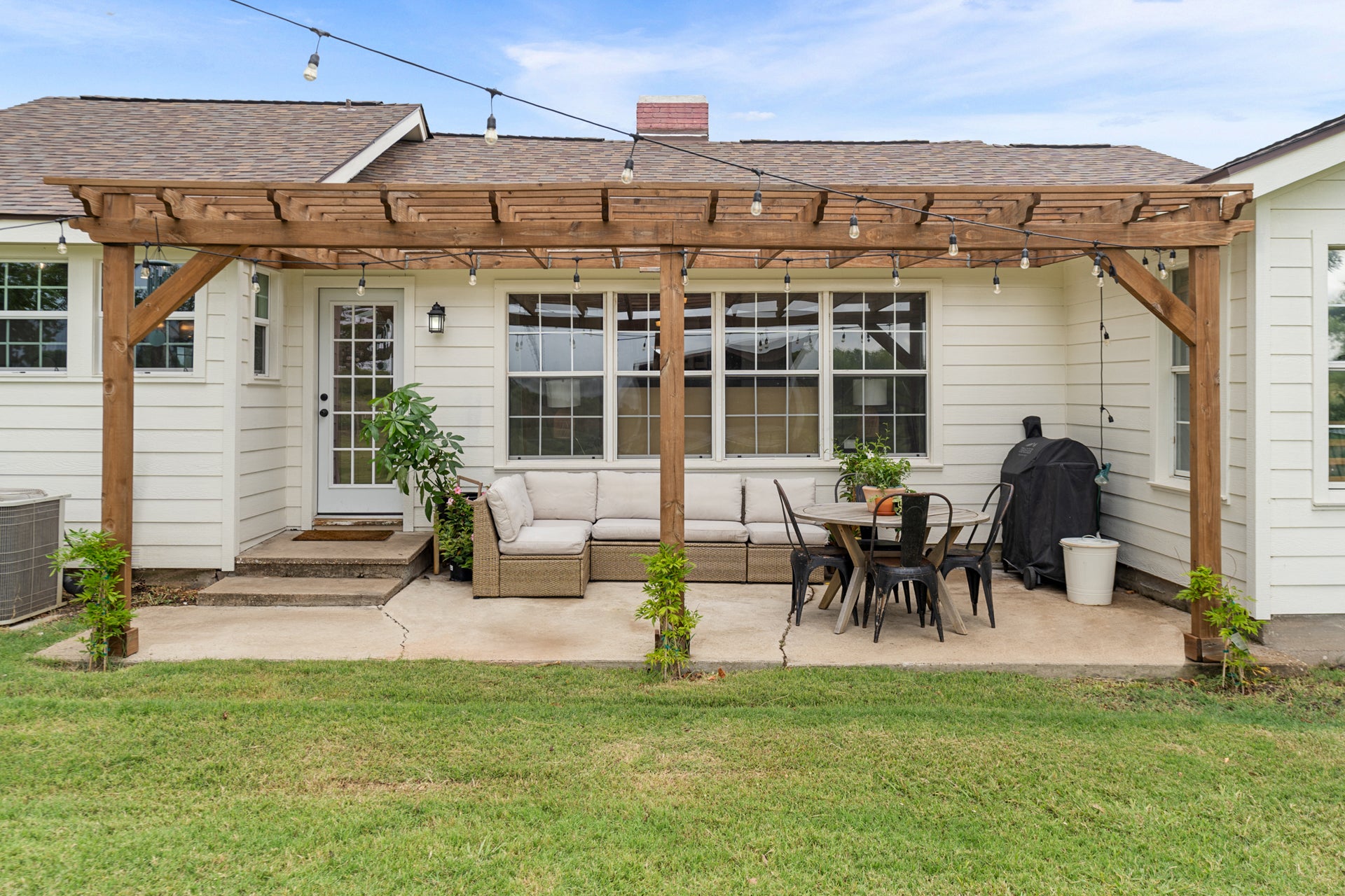 Outdoor patio area with wooden pergola, furniture, grill, and plants.